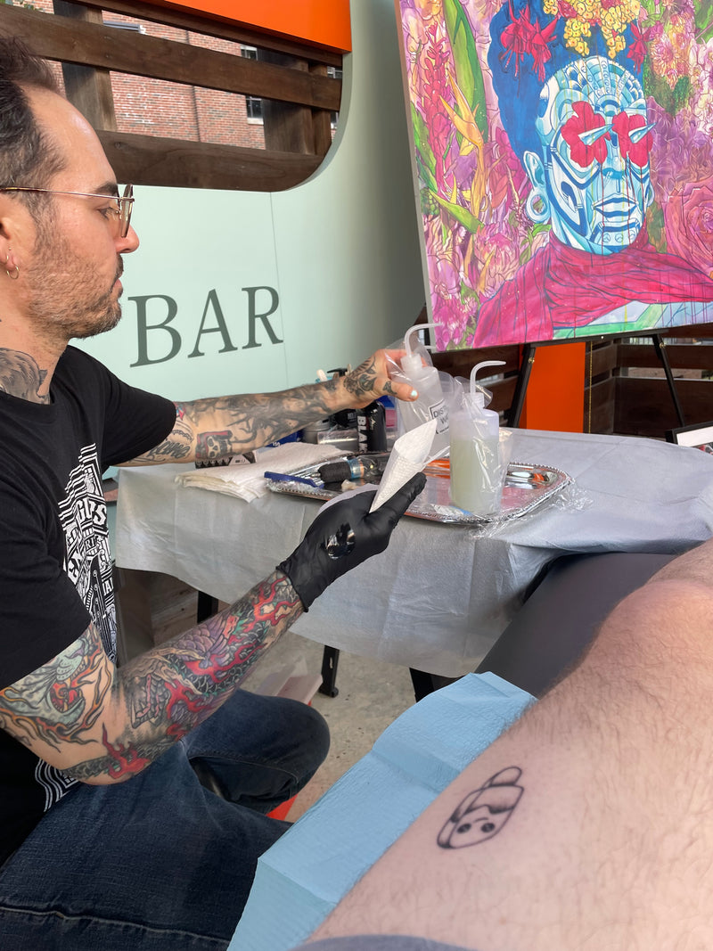 Tattoo artist at work with a colorful tattoo design on a board in the background.